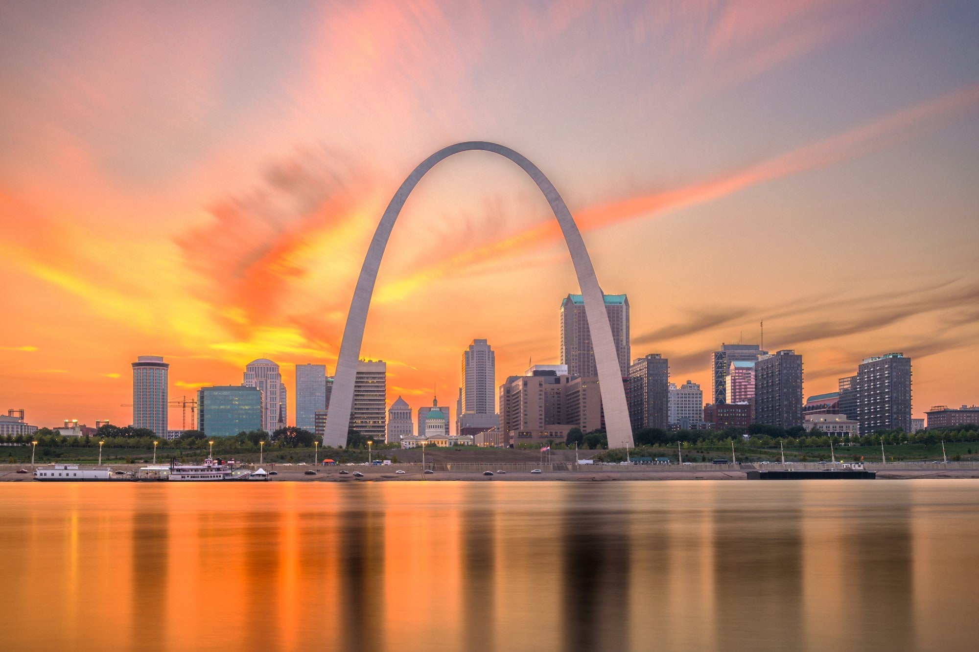 City skyline with the Gateway Arch at sunset, reflecting on a body of water.